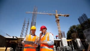 Dois homens vestidos com equipamentos de proteção individual estão em pé, em frente a uma obra enquanto fazem a gestão de obras